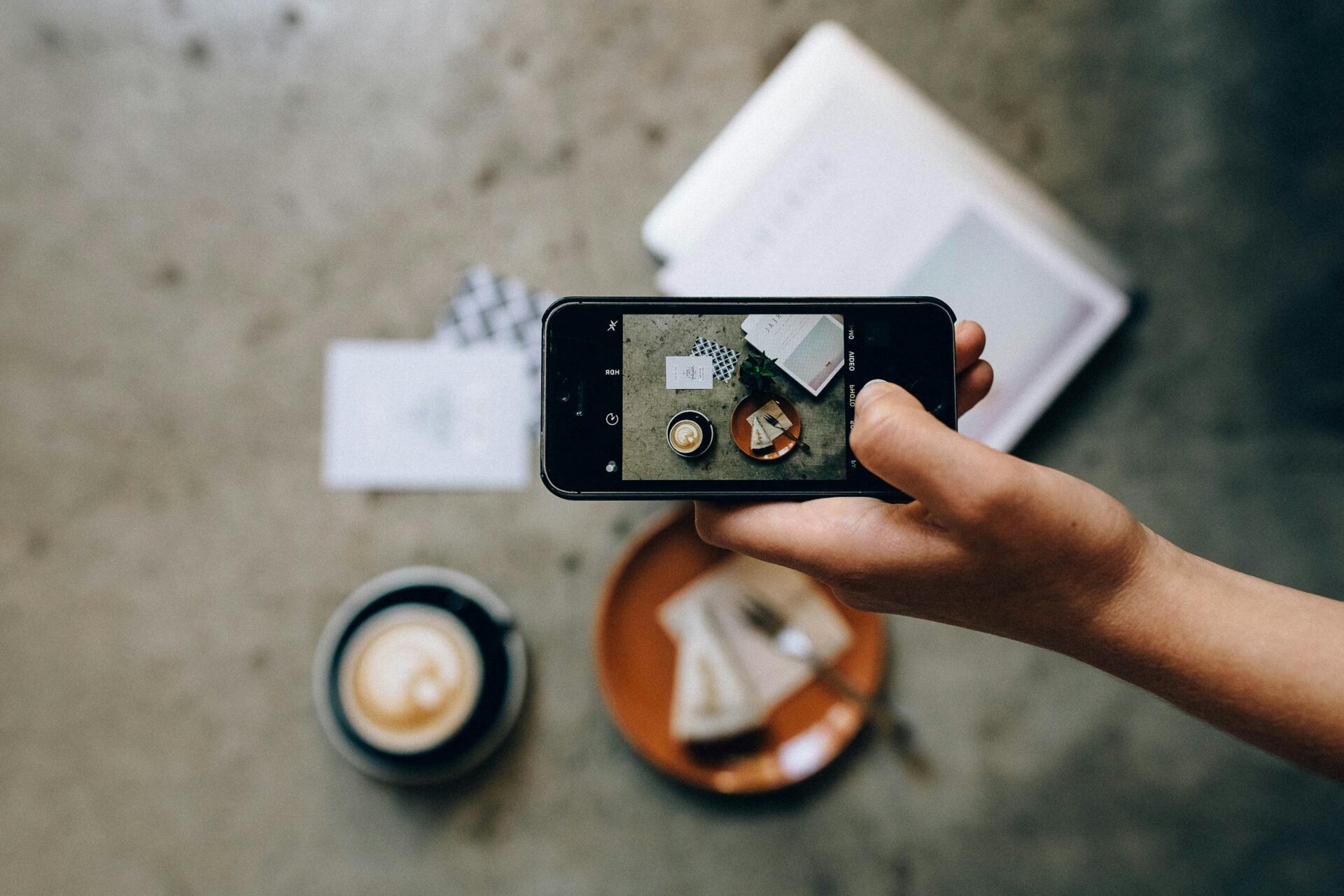 Hand holding smartphone capturing coffee and cake from above.